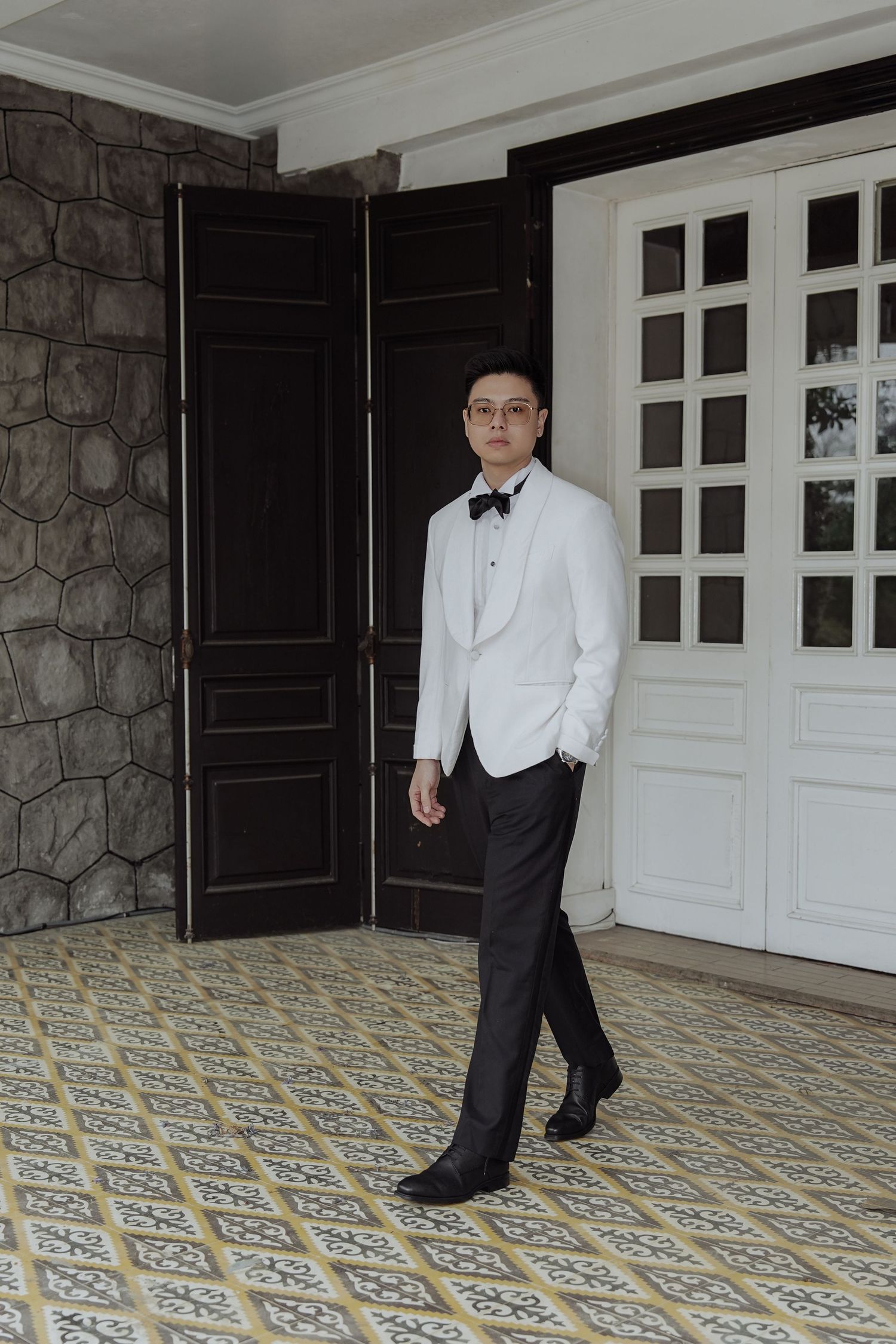 Dapper Asian groom in white tuxedo and black bow tie walking through elegant venue with patterned tile floor.