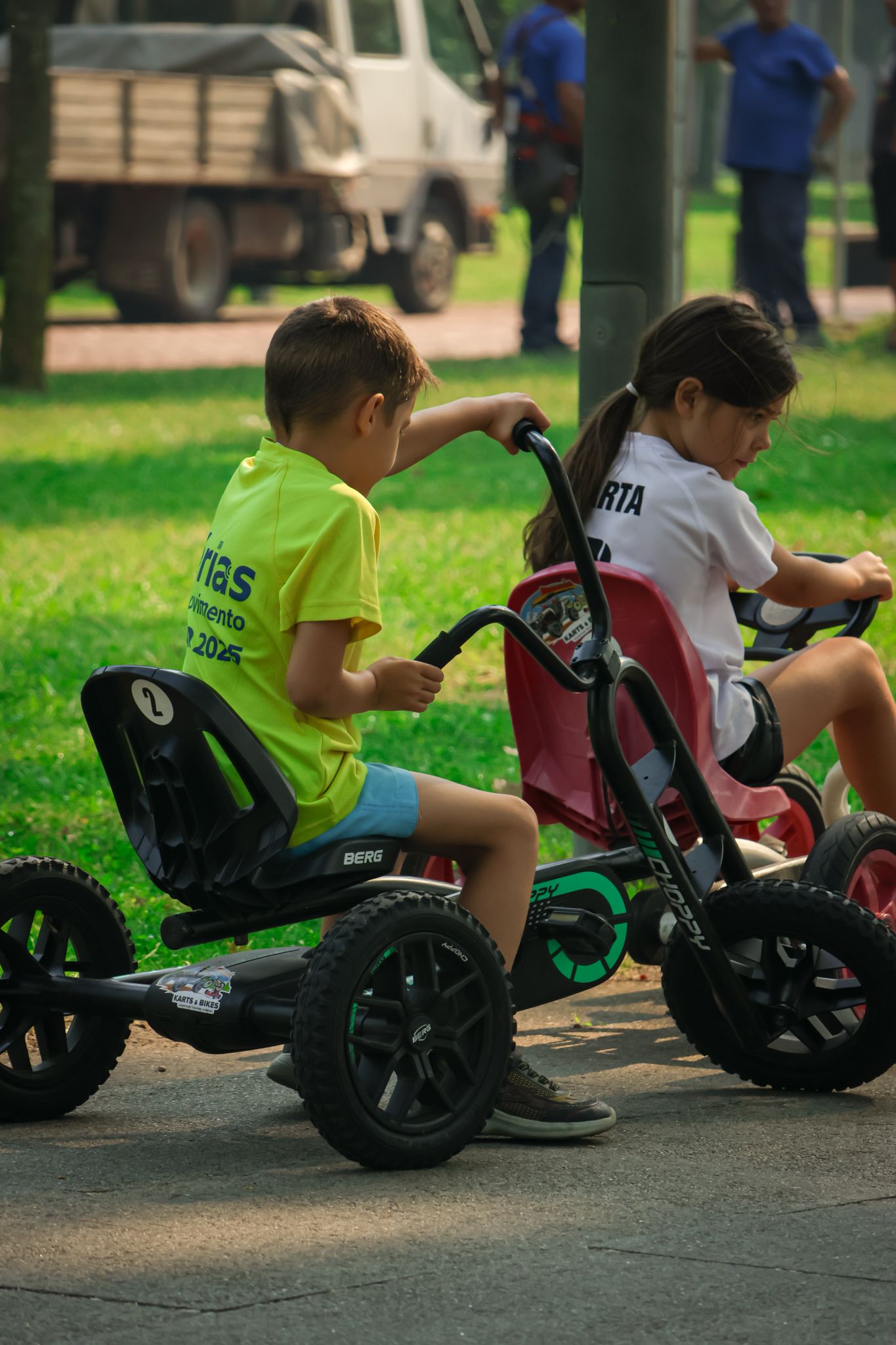 Children sitting on green wheeled riding toys lined up on pavement outdoors.