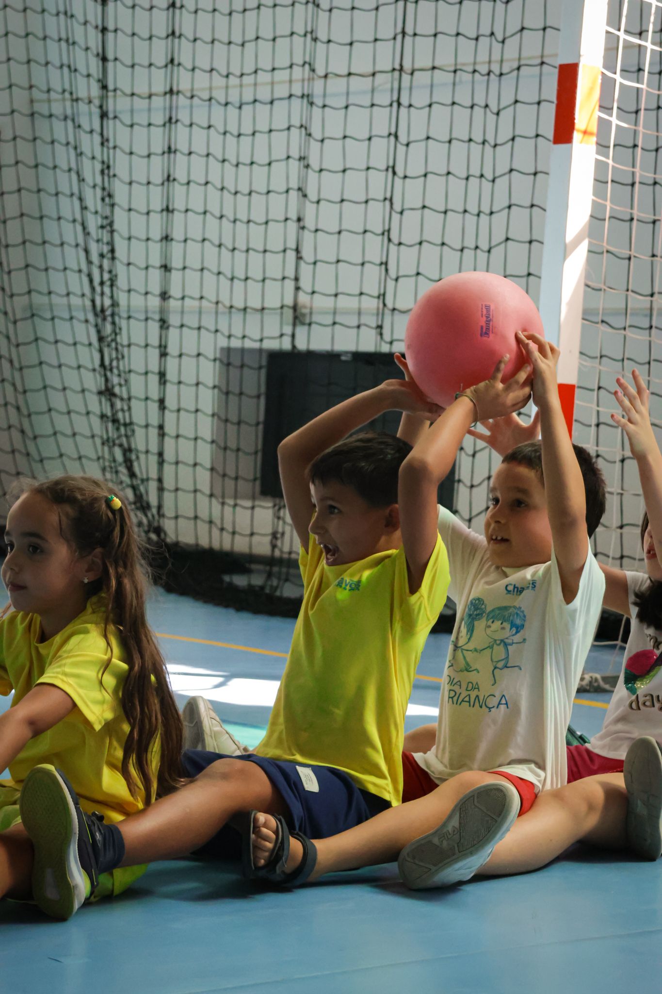 Young athletes in yellow shirts practicing with pink balls while lying on a gym floor.