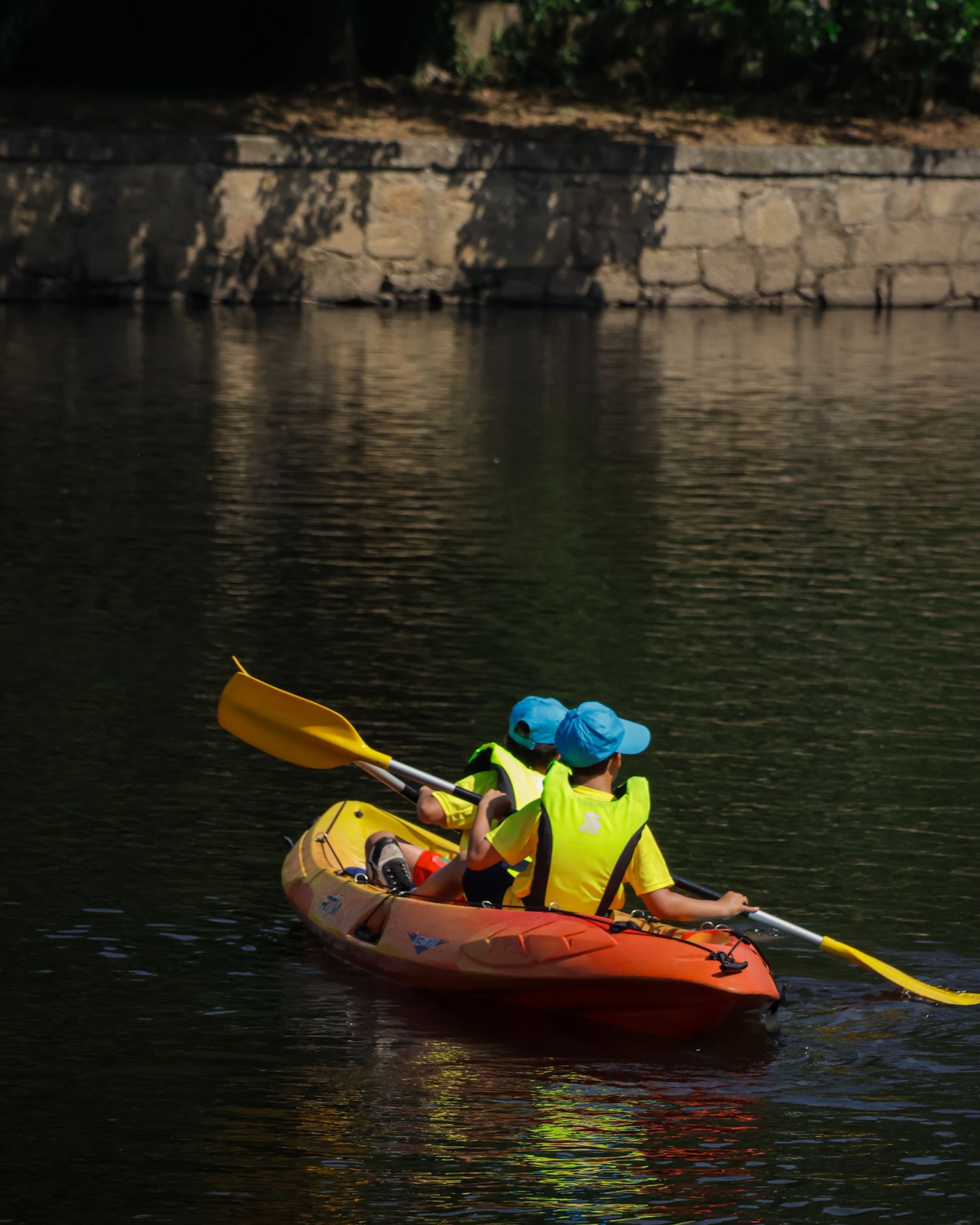 Colorful kayaks glide across dark waters near a stone wall in sequence.