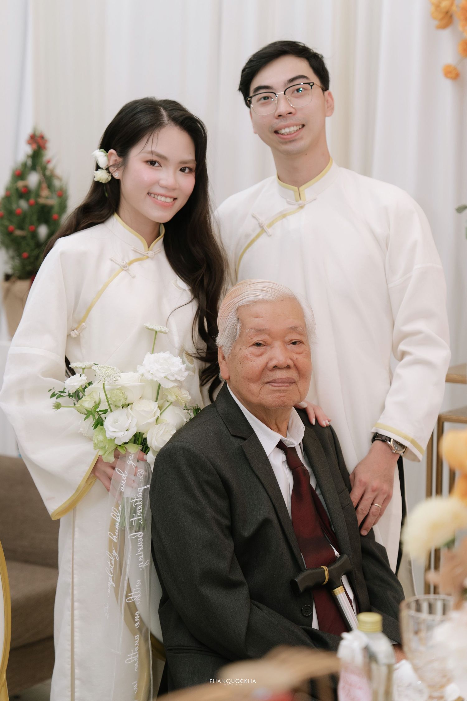 Vietnamese couple in matching white ao dai posing with elderly grandfather at traditional wedding ceremony.
