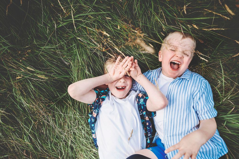 Two children laying in green grass laughing together while wearing blue and white striped clothing.