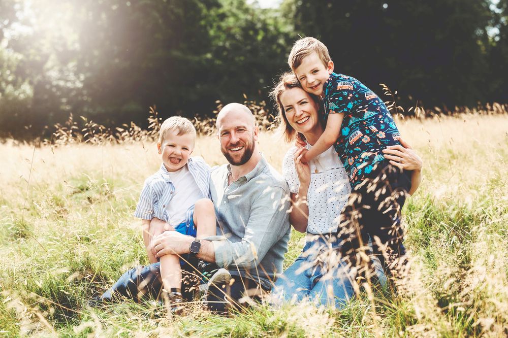 Family members sit together in a sunlit meadow with tall grass and playful expressions during a summer photo session.