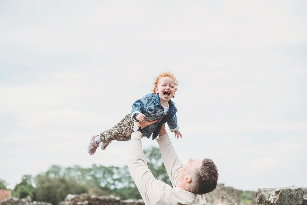 A joyful moment captured outdoors as someone lifts a small child high into the air against a bright sky.