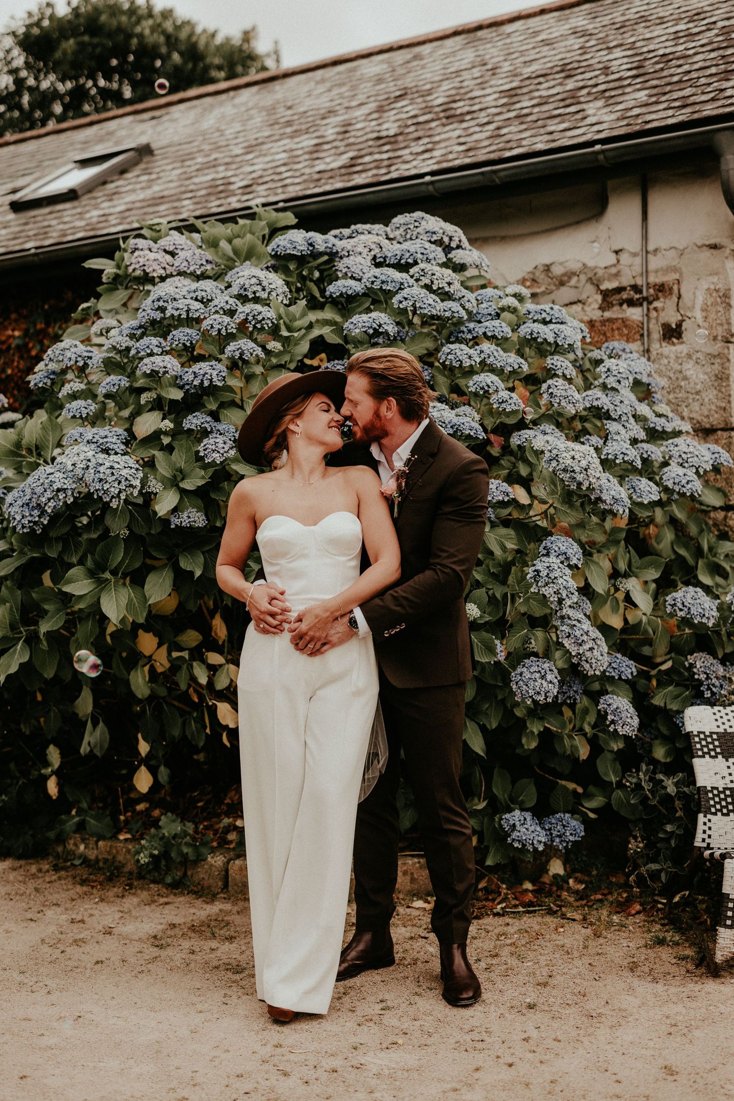 Romantic wedding couple embraces in front of blooming blue hydrangea bush at rustic outdoor venue.