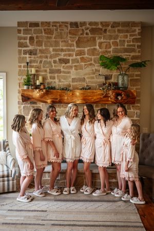 Group wearing matching blush pink robes stand together against rustic stone wall in bridal preparation setting.