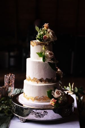 Elegant white wedding cake with gold accents and floral decorations captured in a moody, dark setting.