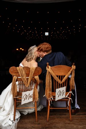 Bride and groom share a romantic kiss while seated on wooden chairs with 'Mr' and 'Mrs' signs at their wedding reception.