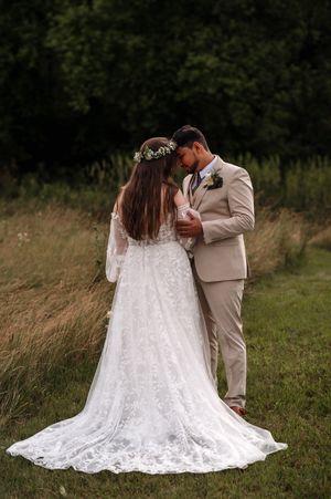 A romantic outdoor wedding photo showing a couple in a flowery meadow, with the bride in a lace gown and flower crown.