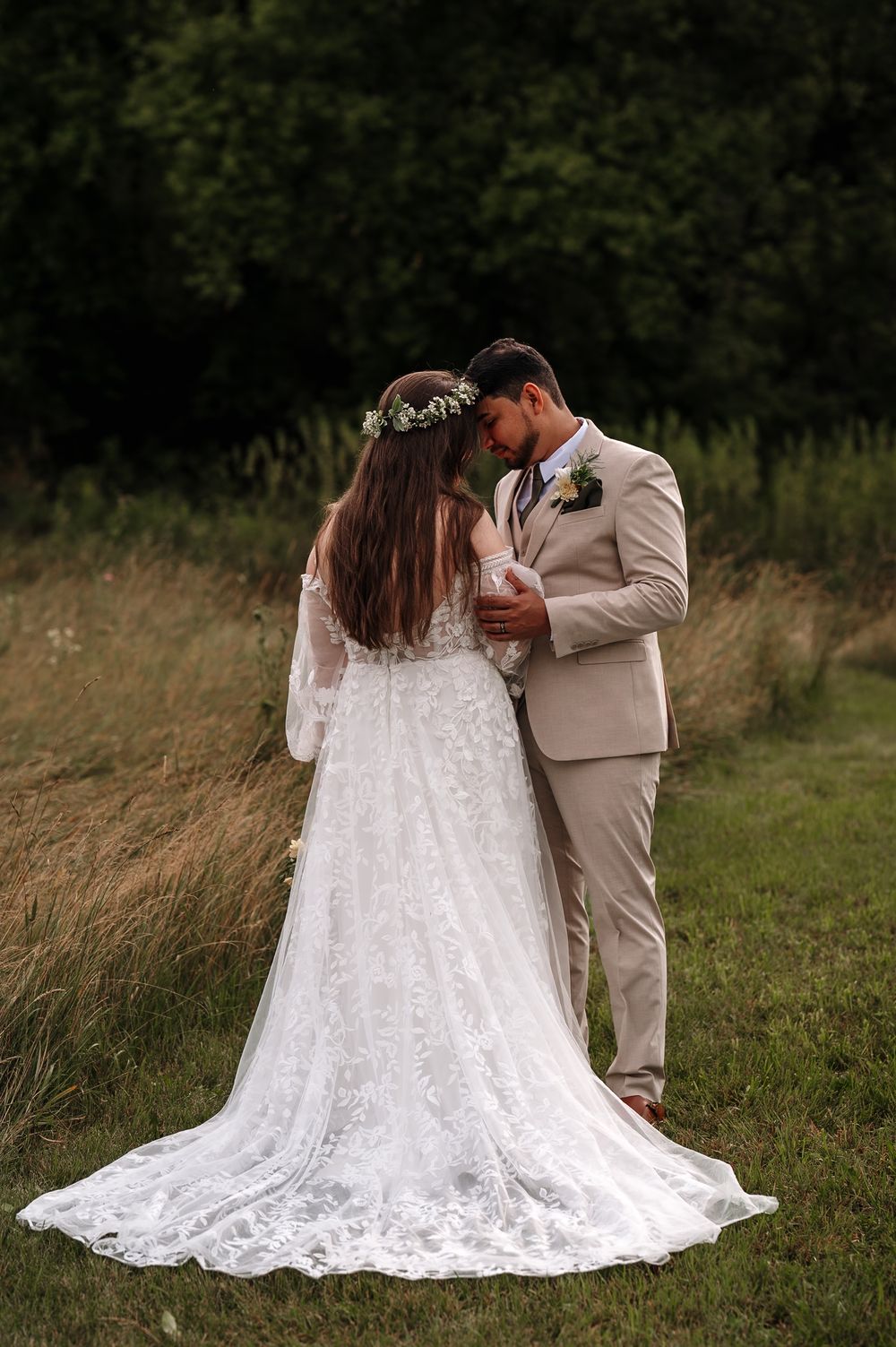 A romantic outdoor wedding photo showing a couple in a flowery meadow, with the bride in a lace gown and flower crown.