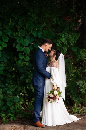 Romantic wedding couple embraces among lush green foliage with bride holding burgundy bouquet.