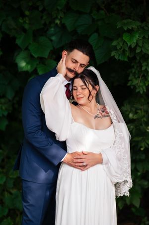 Romantic wedding couple embracing outdoors with bride in flowing white dress against green foliage.
