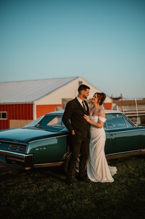 Romantic sunset wedding photo with couple posing beside a classic vintage car near a red barn.