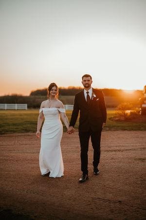 Couple in wedding attire walking together during sunset on a country path.