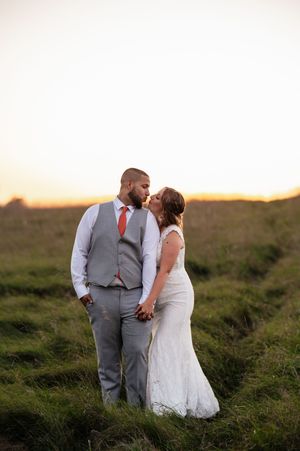 A couple in wedding attire embraces during sunset in a grassy field.