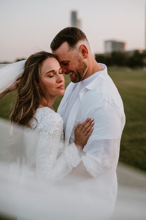Romantic outdoor wedding portrait showing tender moment between couple in white attire against blurred city backdrop.