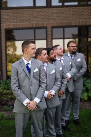 Groomsmen in matching gray suits with blue ties stand in line outdoors near a brick building.