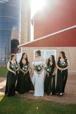 A bridal party in black dresses and white wedding gown pose together in front of rustic farm silos at sunset.