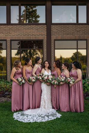 A bridal party wearing dusty rose bridesmaid dresses poses together on grass in front of a brick building at sunset.
