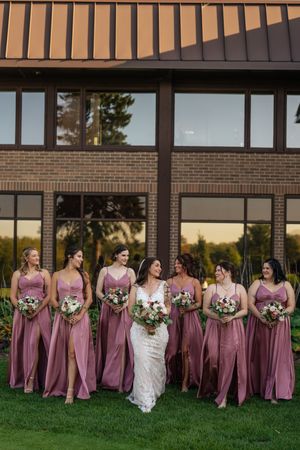 A wedding party in mauve bridesmaid dresses poses outdoors against a brick building at sunset.