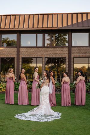 A bride in a white lace gown sits surrounded by bridesmaids wearing dusty rose dresses on a manicured lawn.