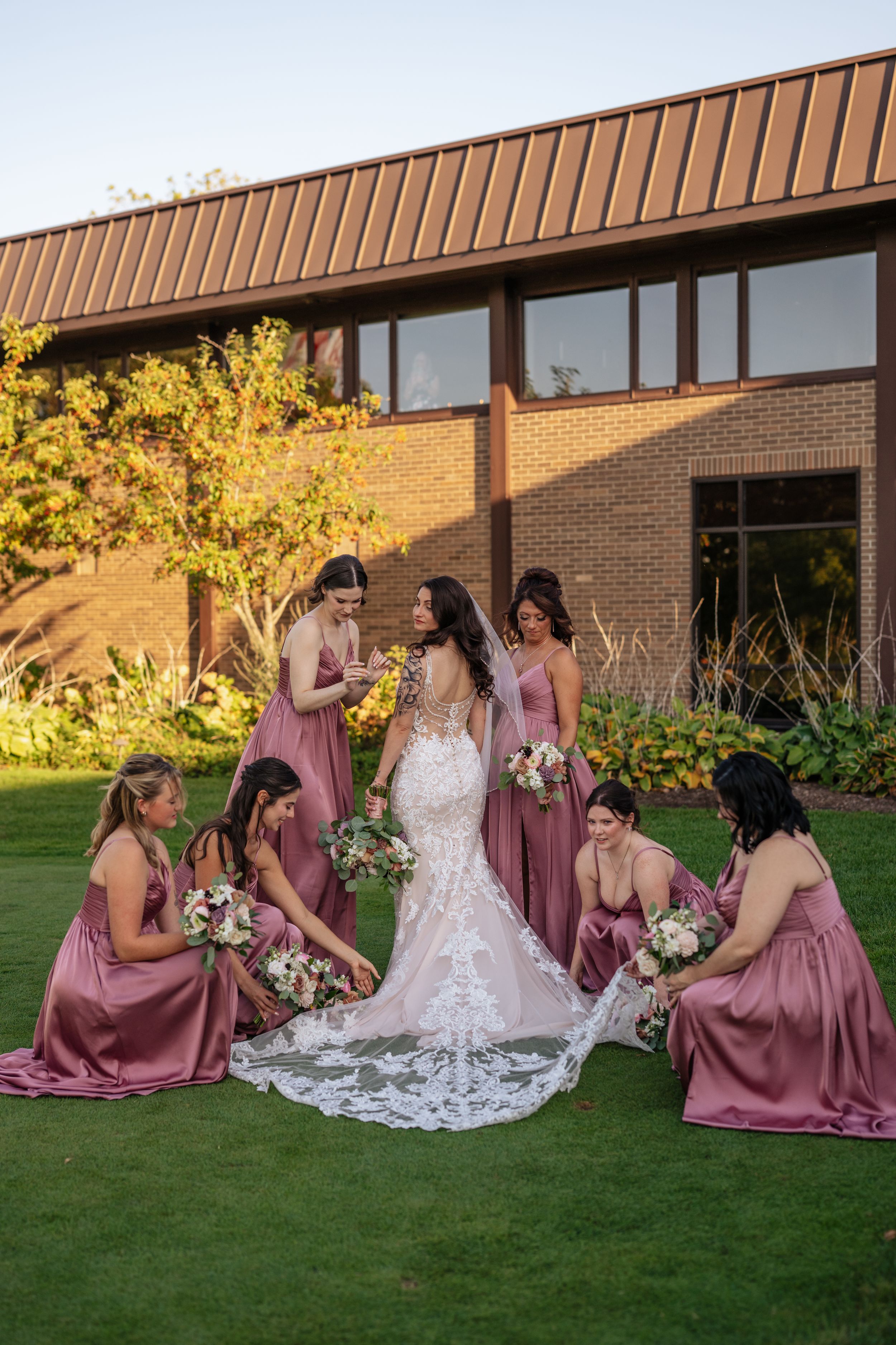 Bridal party in mauve dresses gather around bride in white lace gown on green lawn outside brick building.