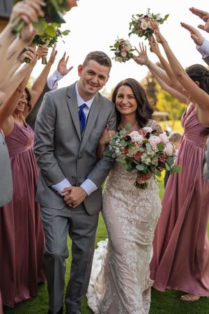 A wedding party celebrates outdoors with bridesmaids in mauve dresses holding bouquets up in the air around the newlyweds.