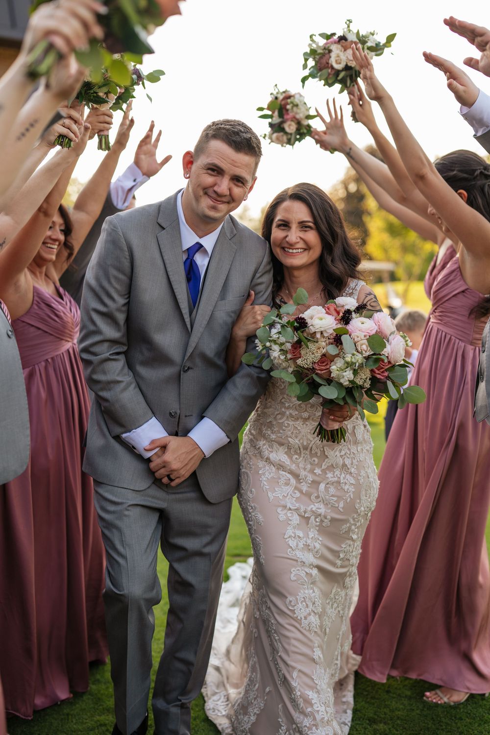 A wedding party celebrates outdoors with bridesmaids in mauve dresses holding bouquets up in the air around the newlyweds.