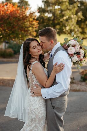 A romantic wedding couple embraces during sunset in an outdoor setting with autumn foliage in the background.