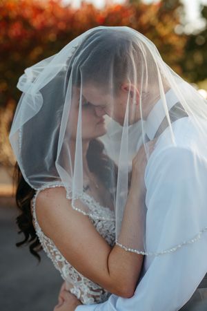 A romantic wedding couple shares an intimate kiss under a delicate white veil with autumn foliage in the background.
