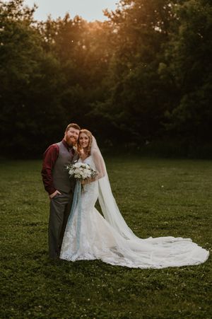 Romantic sunset wedding photos show couple in grassy field with long flowing lace veil trailing behind them.