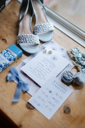 Blue and silver wedding accessories including invitations, ribbons, jewelry and elegant shoes on a wooden surface.