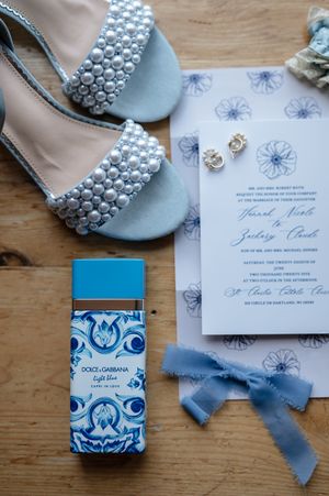 Elegant wedding invitation set with pearl-adorned blue heels and white and blue floral details arranged on wooden surface.