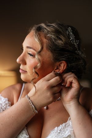 A person wearing bridal jewelry and lace gown is having their earrings put on in a softly lit setting.