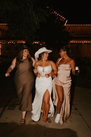 Three women in elegant dresses laugh together while walking at a nighttime event with twinkling lights overhead.