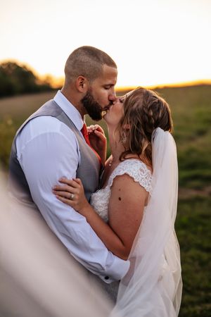 A romantic sunset wedding portrait of a couple kissing outdoors, with the bride in lace and flowing veil.