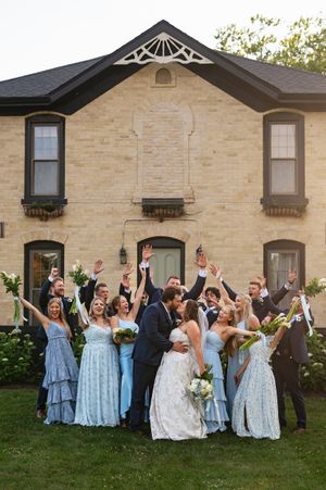 Wedding party in light blue bridesmaid dresses celebrates joyfully on the lawn in front of a stone building.