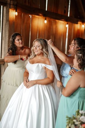 Bride in elegant white off-shoulder ball gown getting help with her veil in rustic wooden venue with warm lighting.