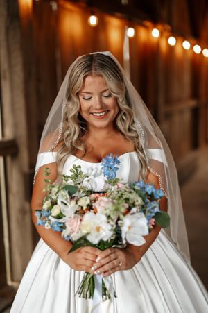 Bride wearing white dress holds beautiful bouquet with pink and white flowers in rustic barn setting with string lights.
