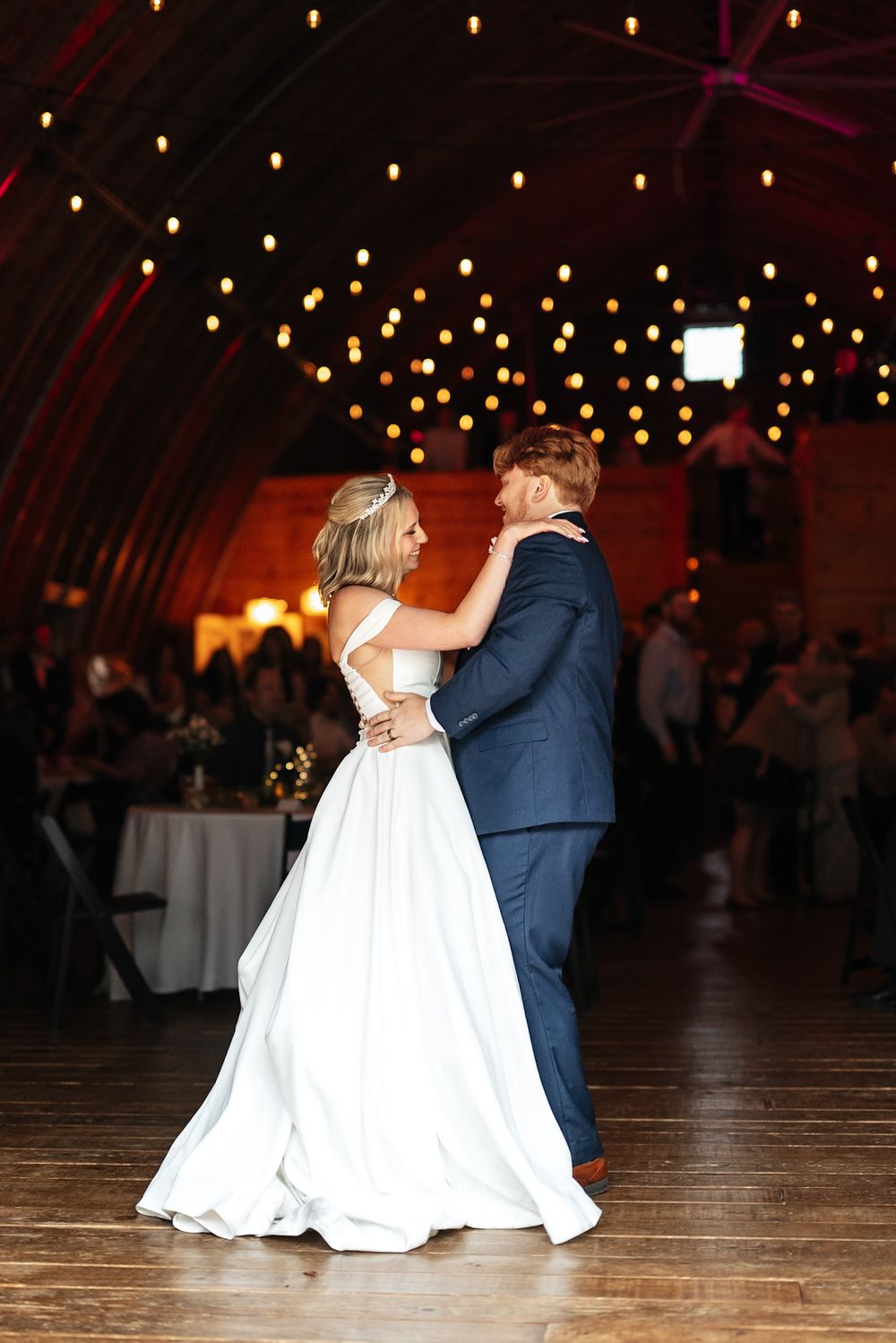 A couple shares their first dance at a rustic wedding reception under twinkling lights.
