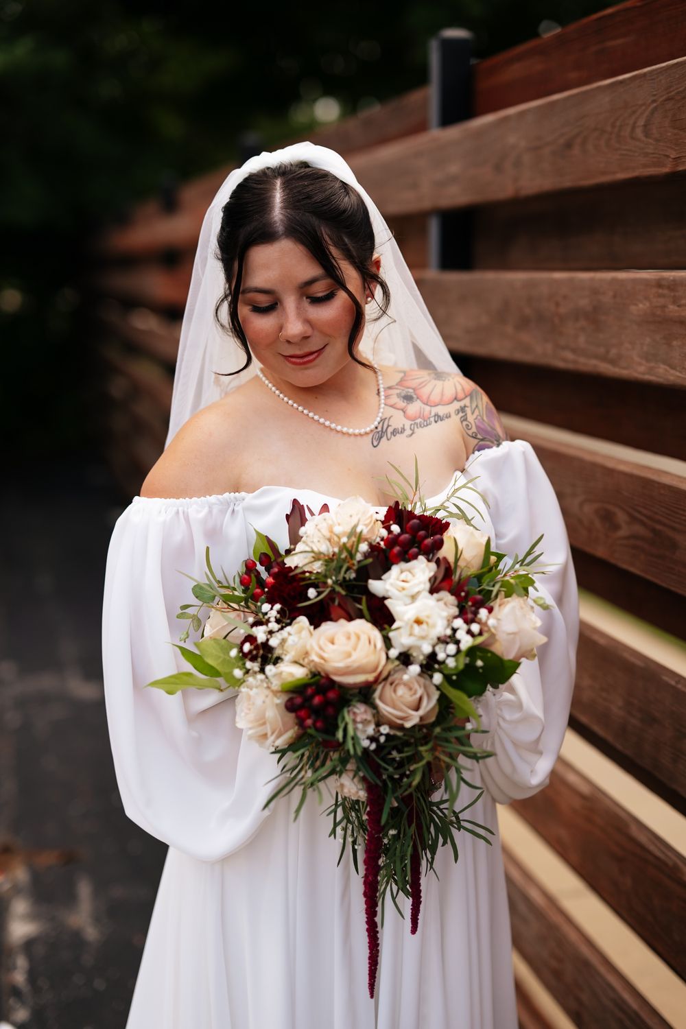 A woman in a white dress holds a beautiful burgundy and blush wedding bouquet beside wooden stairs.