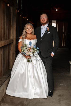 Wedding couple poses in rustic barn setting in formal attire with bride holding white floral bouquet.