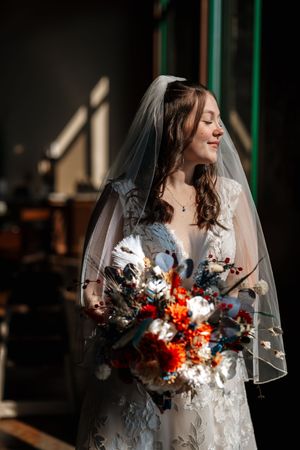 A bride in a white lace gown holds a vibrant autumn bouquet with orange and burgundy flowers against a dark background.