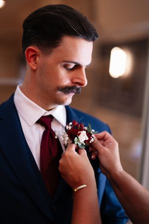 Close-up view of someone helping to pin a dark red boutonniere with white accents onto a navy blue suit jacket.