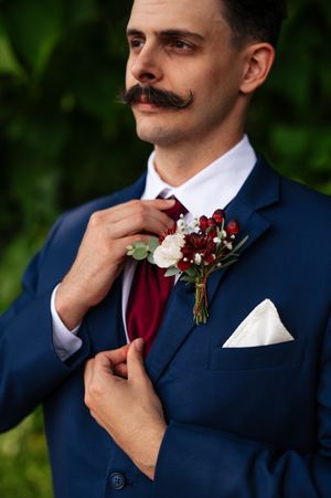 Stylish groom in navy suit with handlebar mustache and burgundy tie adjusts collar in outdoor portrait series.