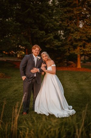 A romantic wedding photo of a couple standing in a grassy field at golden hour with trees in the background.