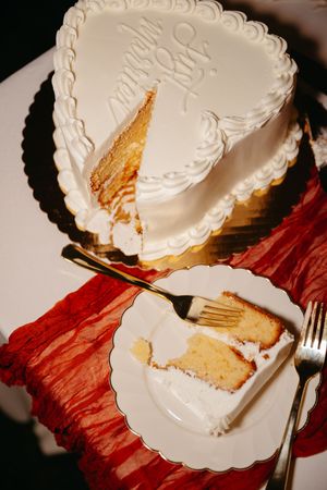 A white heart-shaped wedding cake with one slice removed sits on a plate with red decor.