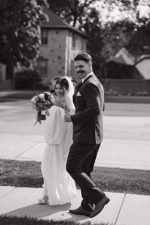 A black and white photo of a bride and groom walking together outdoors, with the bride holding a floral bouquet.