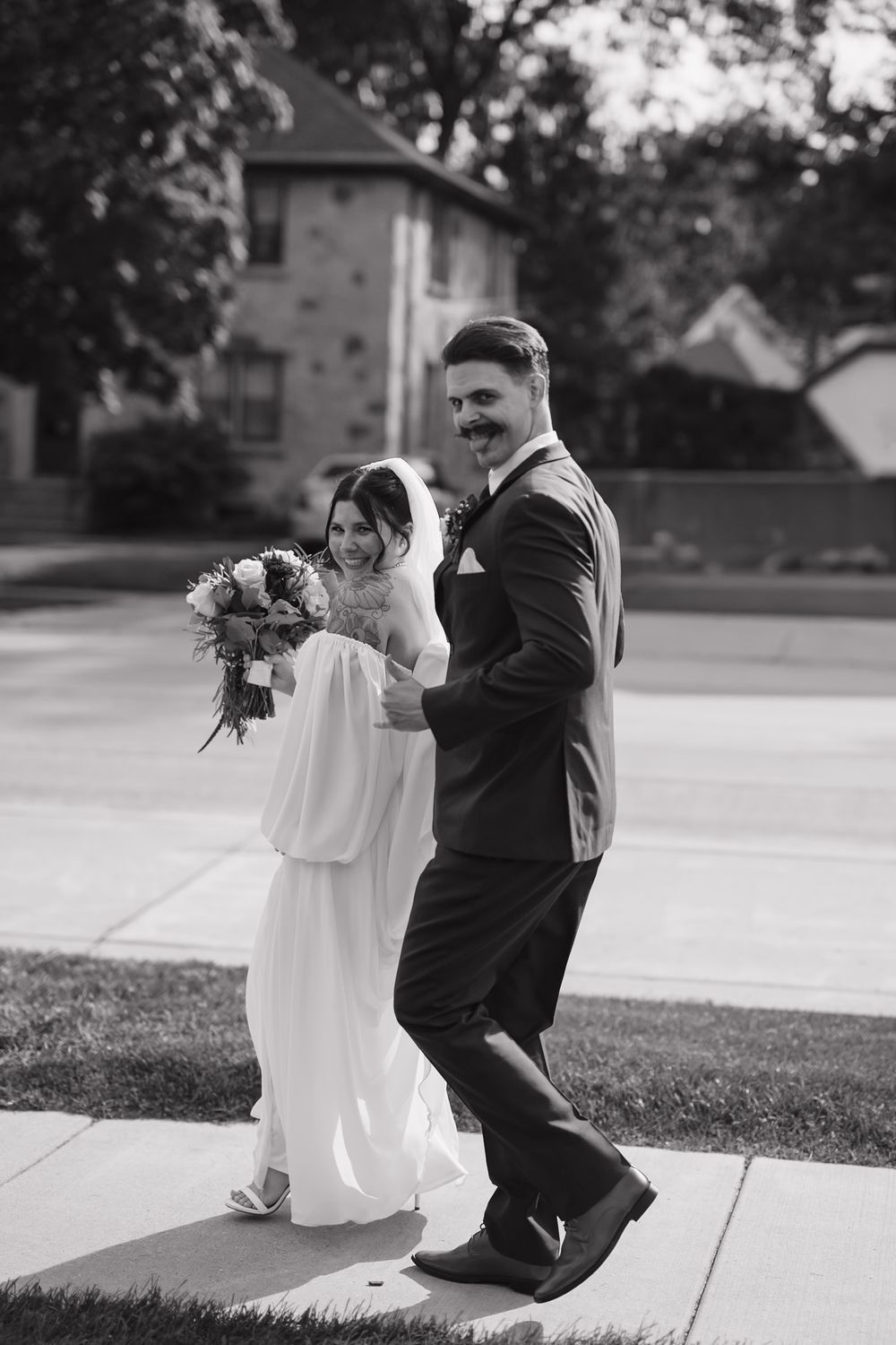 A black and white photo of a bride and groom walking together outdoors, with the bride holding a floral bouquet.