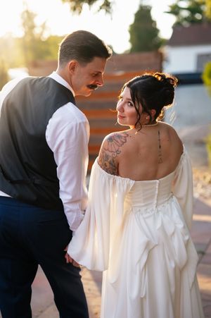 A bride in a flowy white off-shoulder dress and groom in dark suit share intimate moments during their outdoor wedding photoshoot.